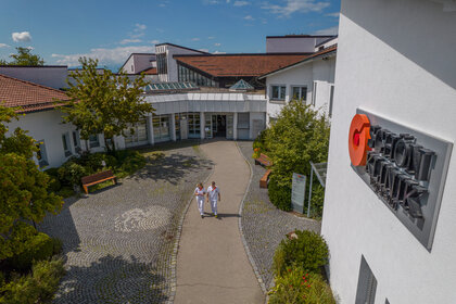 Eingangsbereich der Schön Klinik Vogtareuth mit weißem Gebäude, Glasdach, Vorhof, Bänken und zwei Mitarbeitenden im Freien