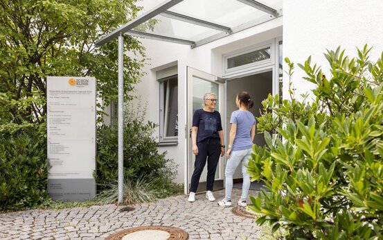 Zwei Personen vor Eingang, weißes Gebäude mit Glasvordach und offenem Zugang