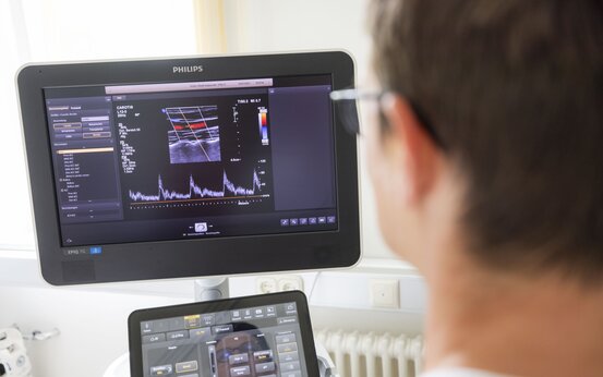 Arzt betrachtet Ultraschallmonitor mit farbiger Doppleraufnahme zur Gefäßdiagnostik in heller Praxis