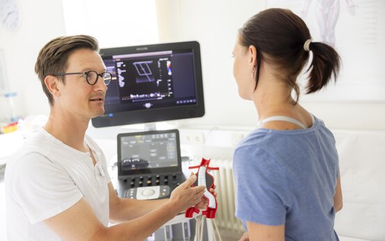 Arzt erklärt Patientin mit Blutgefäßmodell die Ultraschalldiagnostik, Ultraschallgerät im Hintergrund