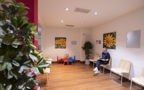 Helles Wartezimmer mit Holzstühlen, älterer Person beim Lesen, Pflanzen und Kinderspielecke mit bunten Möbeln