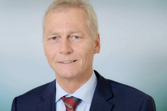 Mann mit kurz blond-grauem Haar, dunkelblauem Anzug, hellblauem Hemd und rot-blauer Krawatte vor hellem Hintergrund, lächelt