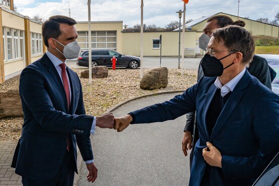 Zwei Männer in Anzügen mit medizinischen Masken begrüßen sich draußen vor einem Gebäude mit Faustgruß, weitere Person im Hintergrund