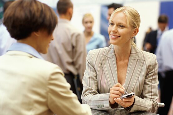 Zwei Personen in hellen Blazern sitzen sich an einem runden Tisch gegenüber, eine Person hält ein Smartphone, beide lächeln sich an