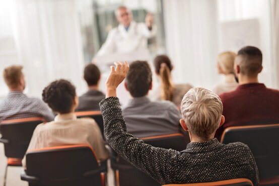 Person hebt im Publikum die Hand, Arzt im weißen Kittel hält Vortrag für Patienten und Angehörige im Seminarraum