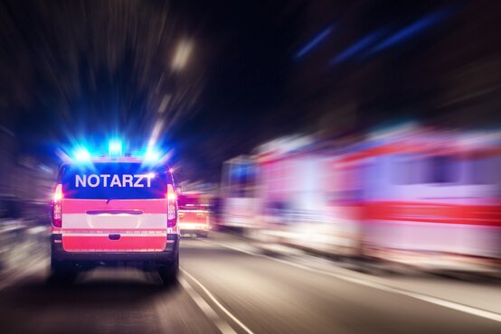 Rettungswagen mit der Aufschrift „Notarzt“ und Blaulicht fährt nachts auf zweispuriger Straße, weitere Einsatzfahrzeuge am Rand