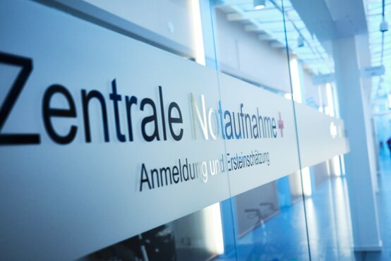 Heller Klinikflur mit Glaswand, Aufschrift „Zentrale Notaufnahme + Anmeldung und Ersteinschätzung“