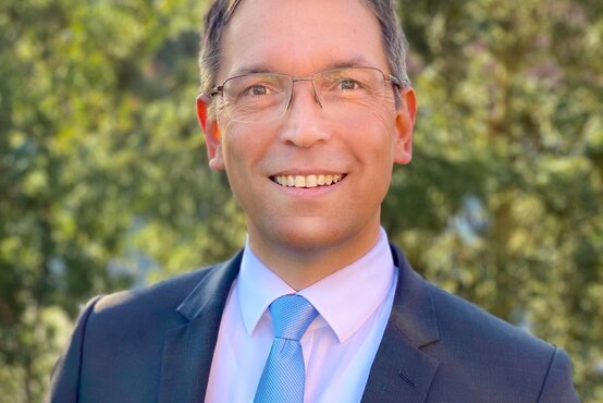 Mann mit kurzem braunem Haar und Brille, dunkelblauer Anzug, weißes Hemd, hellblaue Krawatte, lächelt im Freien vor grünem Hintergrund