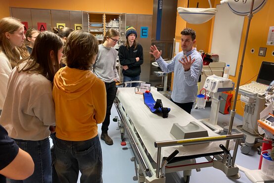 Mehrere Jugendliche im Schockraum um ein Untersuchungsbett, Mann erklärt medizinische Geräte und Hilfsmittel, Präventionsprogramm