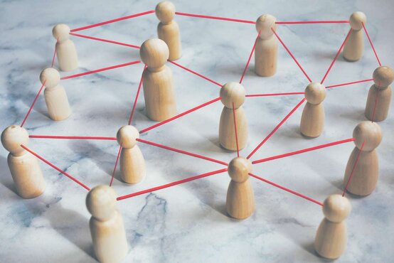 Holzfiguren auf Marmorfläche sind mit roten Fäden zu einem Netzwerk verbunden, Symbol für Zusammenarbeit im Parkinson-Netzwerk
