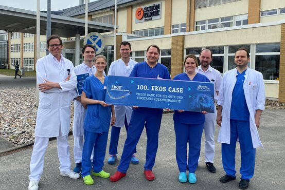 Acht Fachkräfte der Schön Klinik Neustadt halten vor dem Klinikgebäude ein blaues Schild „100. EKOS CASE“