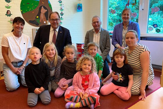 Gruppenfoto von fünf Erwachsenen und sechs Kindern in bunter Kleidung in einem freundlichen Kindergartenraum mit gemalten Wandbildern