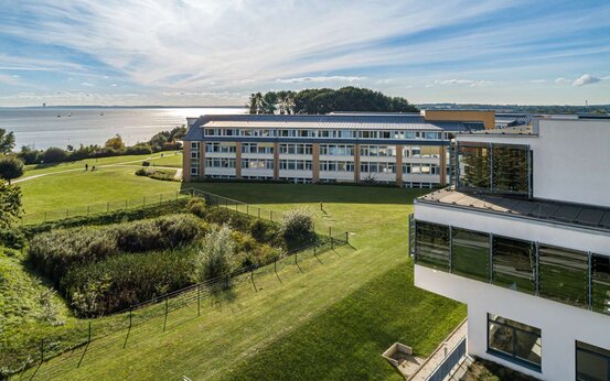 Luftaufnahme eines der Schön Klinik Neustadt mit großen Fenstern, grüner Wiese und Meer im Hintergrund