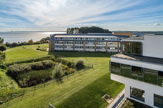 Luftaufnahme eines der Schön Klinik Neustadt mit großen Fenstern, grüner Wiese und Meer im Hintergrund