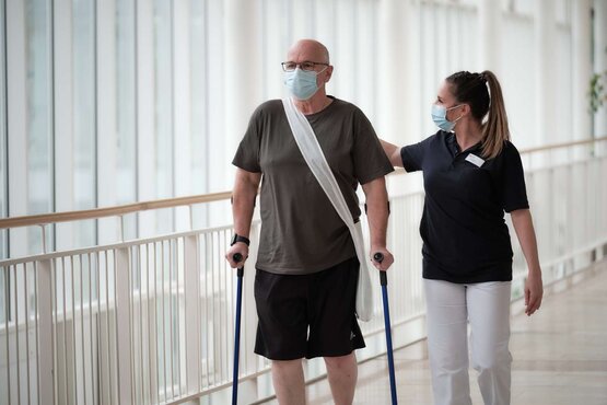 Mann mit Umhängetasche und Gehstützen läuft neben Therapeutin, beide mit Mund-Nasen-Masken im Klinikflur