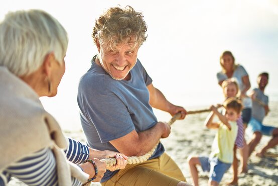 Familie mit Großeltern, Eltern und Kindern beim Tauziehen am Sandstrand, alle lachen und genießen das sonnige Wetter