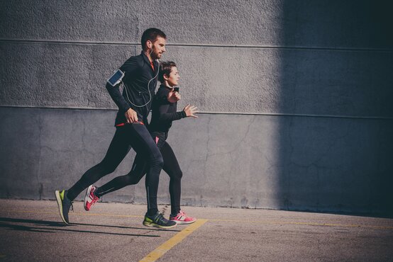 Junges Paar in dunkler Sportkleidung joggt nebeneinander mit Kopfhörern vor grauer Wand