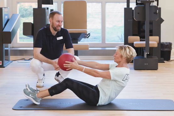Frau trainiert auf Gymnastikmatte im Fitnessstudio, hält roten Medizinball, Therapeut beobachtet sie, Fitnessgeräte im Hintergrund