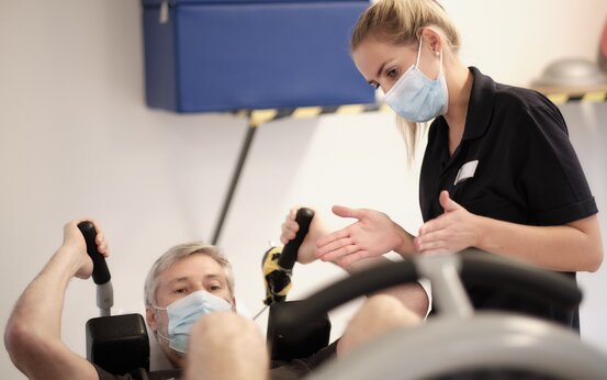 Mann mit grauen Haaren sitzt an Fitnessgerät und hält schwarze Griffe, Therapeut steht daneben und unterstützt