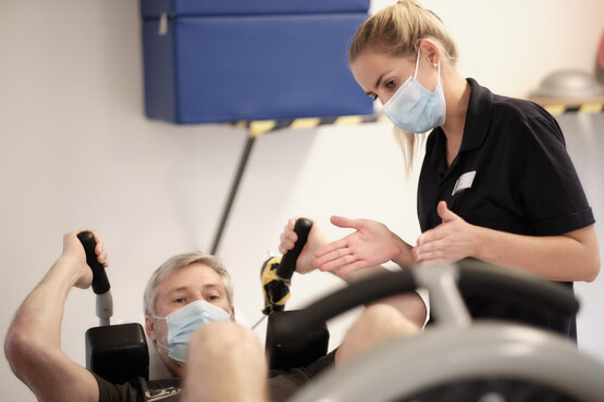 Mann mit grauen Haaren sitzt an Fitnessgerät und hält schwarze Griffe, Therapeut steht daneben und unterstützt