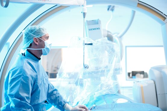 Ärzteteam in blauer OP-Kleidung und Mundschutz arbeitet im modernen Hybrid-Operationssaal mit großem Monitor und medizinischen Geräten