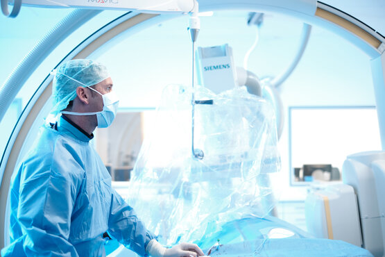 Ärzteteam in blauer OP-Kleidung und Mundschutz arbeitet im modernen Hybrid-Operationssaal mit großem Monitor und medizinischen Geräten