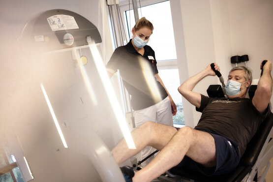Patient mit Maske trainiert an Fitnessgerät, Therapeutin mit Maske steht daneben in hellem Therapieraum