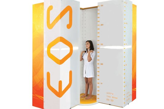 Frau im weißen Kittel steht barfuß auf gelber Plattform in weißer EOS-Röntgenkabine mit oranger Aufschrift und Messskalen