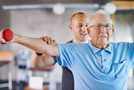 Mann mit Brille und hellblauem Poloshirt hebt roten Hantelarm, Physiotherapeut unterstützt an Schulter