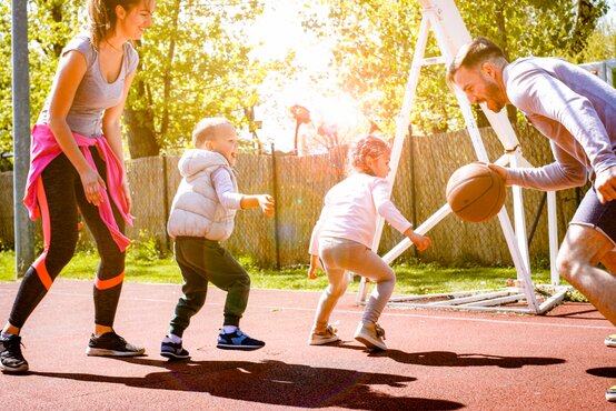 Familie spielt Basketball auf Outdoor-Sportplatz, zwei Erwachsene und zwei Kinder