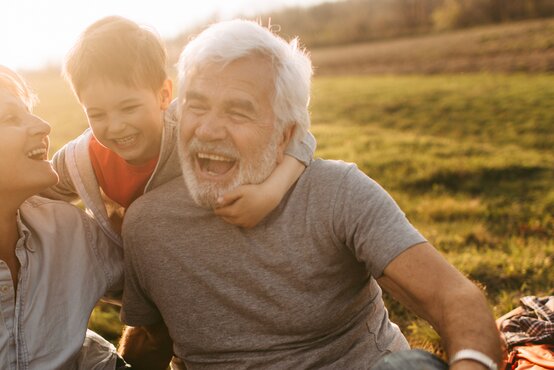 Älteres Paar sitzt mit Kind auf Picknickdecke im Park, alle lachen und genießen gemeinsame Zeit
