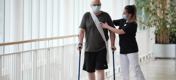 Älterer Mann mit Umhängetasche und Gehstützen läuft neben Frau, beide mit Mund-Nasen-Masken im Klinikflur