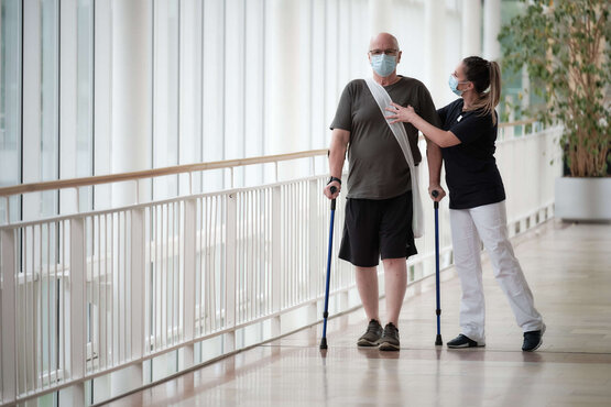 Mann mit Gehhilfen und Schön Klinik Patiententasche wird von Pflegekraft in hellem Flur unterstützt