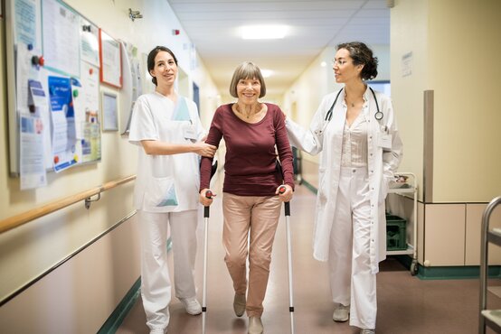 Ältere Frau mit Krücken wird im Krankenhausflur von Ärztin und Pflegekraft unterstützt