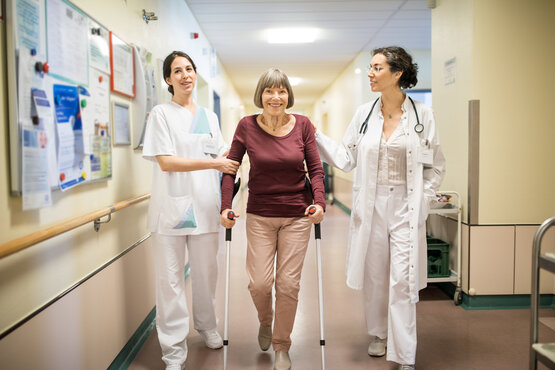 Ältere Frau mit Krücken wird im Krankenhausflur von Ärztin und Pflegekraft unterstützt