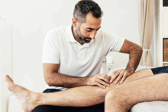 Physiotherapeut behandelt rechtes Bein eines Patienten auf Liege, manuelle Therapie in heller Praxisumgebung