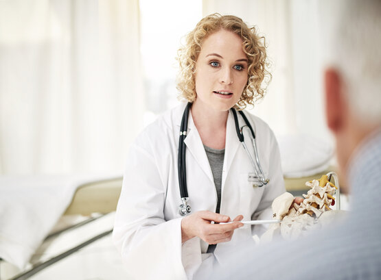 Ärztin mit lockigen blonden Haaren zeigt mit Stift auf Wirbelsäulenmodell, spricht mit Person im hellen Arztzimmer