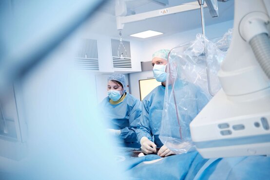 Zwei Personen in blauen OP-Kitteln, Hauben und Masken stehen im hellen Operationssaal neben medizinischem Gerät mit transparenter Folie
