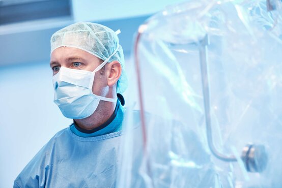 Arzt in blauer OP-Kleidung, Haube und Mundschutz steht im hellen Operationssaal 