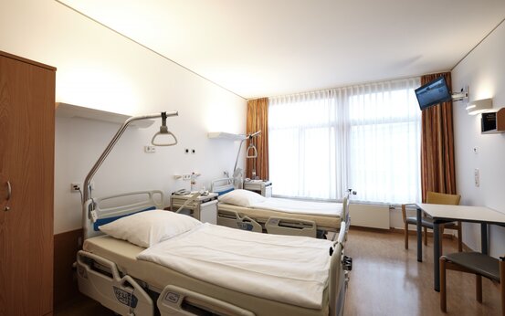Patientenzimmer mit Holzschrank, Tisch, Stühlen und Fenster mit Gardinen