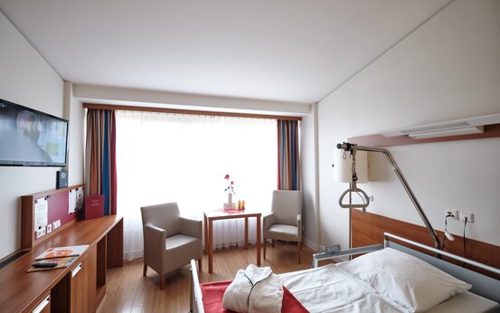 Helles, modern eingerichtetes Patientenzimmer mit Pflegebett, Sitzgruppe, Schreibtisch und Flachbildfernseher