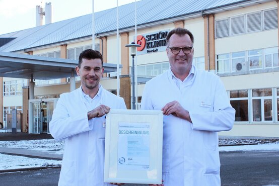 Zwei Ärzte im weißen Kittel halten eine gerahmte Bescheinigung vor dem Eingang der Schön Klinik Neustadt