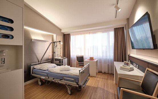 Modernes, helles Patientenzimmer mit großem Fenster, Pflegebett, Nachtschrank, Flachbildfernseher, Stuhl und Holzfußboden