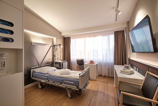 Modernes, helles Patientenzimmer mit großem Fenster, Pflegebett, Nachtschrank, Flachbildfernseher, Stuhl und Holzfußboden