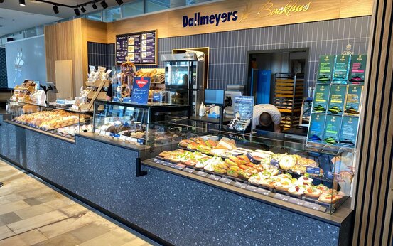Bäckereitheke mit Glasvitrine voller Backwaren, Mitarbeiterin dahinter, Logo „Dallmeyers Backhus“ an der Wand