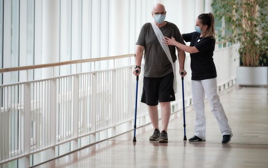 Älterer Mann mit Umhängetasche läuft mit Krücken und wird von Physiotherapeutin unterstützt