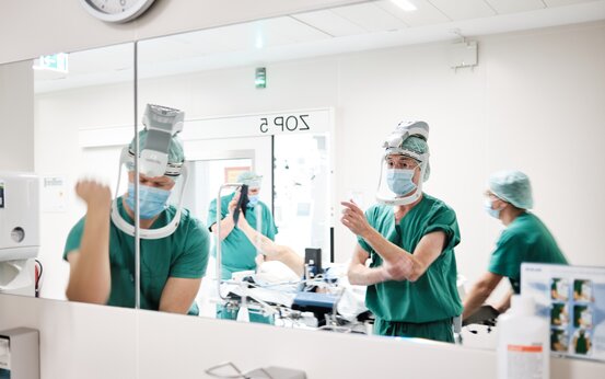 Personen in grüner OP-Kleidung, Haube, Maske und Visier stehen im OP-Raum, Patient auf Liege im Hintergrund