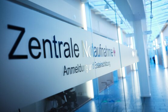 Heller Klinikflur mit Glaswand, Aufschrift „Zentrale Notaufnahme + Anmeldung und Ersteinschätzung“