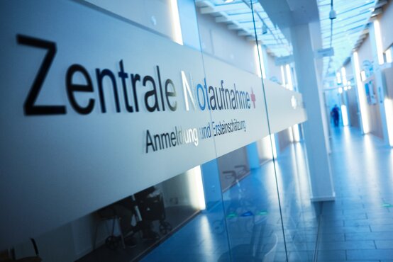 Heller Klinikflur mit Glaswand, Aufschrift „Zentrale Notaufnahme + Anmeldung und Ersteinschätzung“