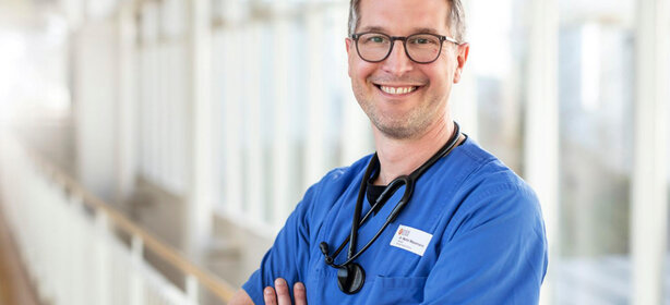 Person in blauer Arbeitskleidung mit Stethoskop im modernen, hellen Klinikflur